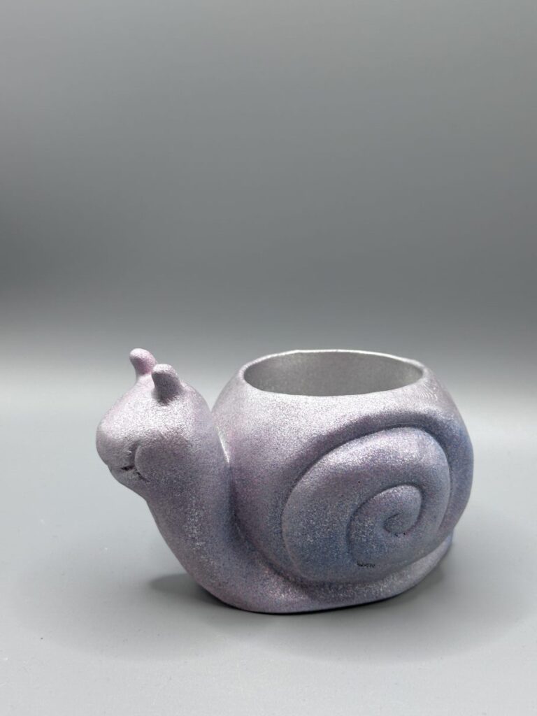 A small, smiling snail-shaped plant pot with a shimmering iridescent lavender and light blue metallic finish against a grey background.