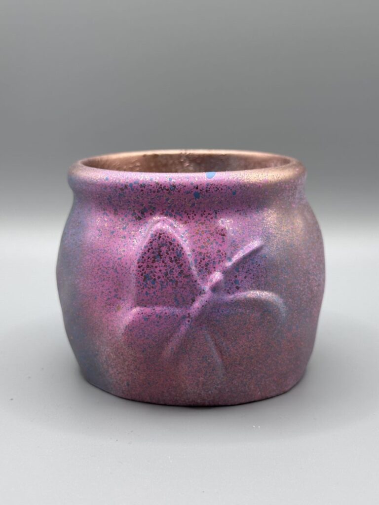 A small, rounded pot with an embossed butterfly, featuring a vibrant pink and purple speckled finish with blue accents and a bronze metallic rim.