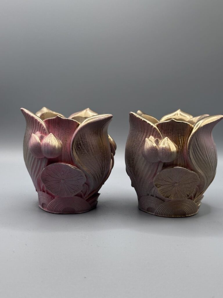 Two small, ornate planters shaped like lotus flowers with a textured rose and gold metallic gradient finish, displayed on a grey surface.