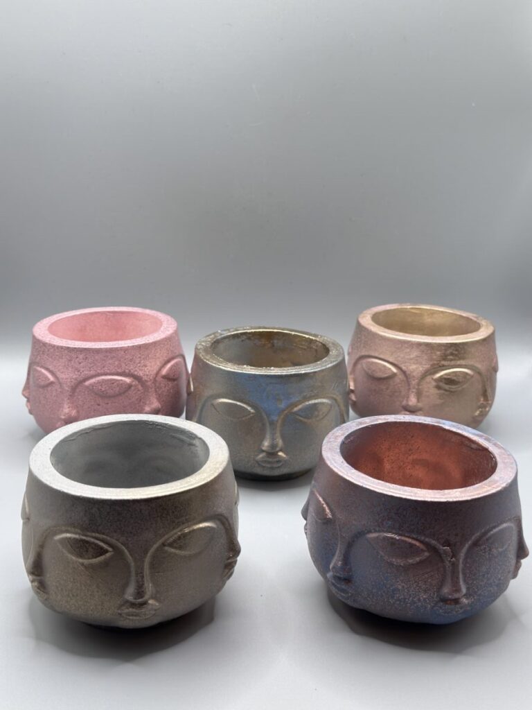 A collection of five small, round planters featuring embossed minimalist faces, finished in various metallic gradients including pink, blue, gold, and pewter.