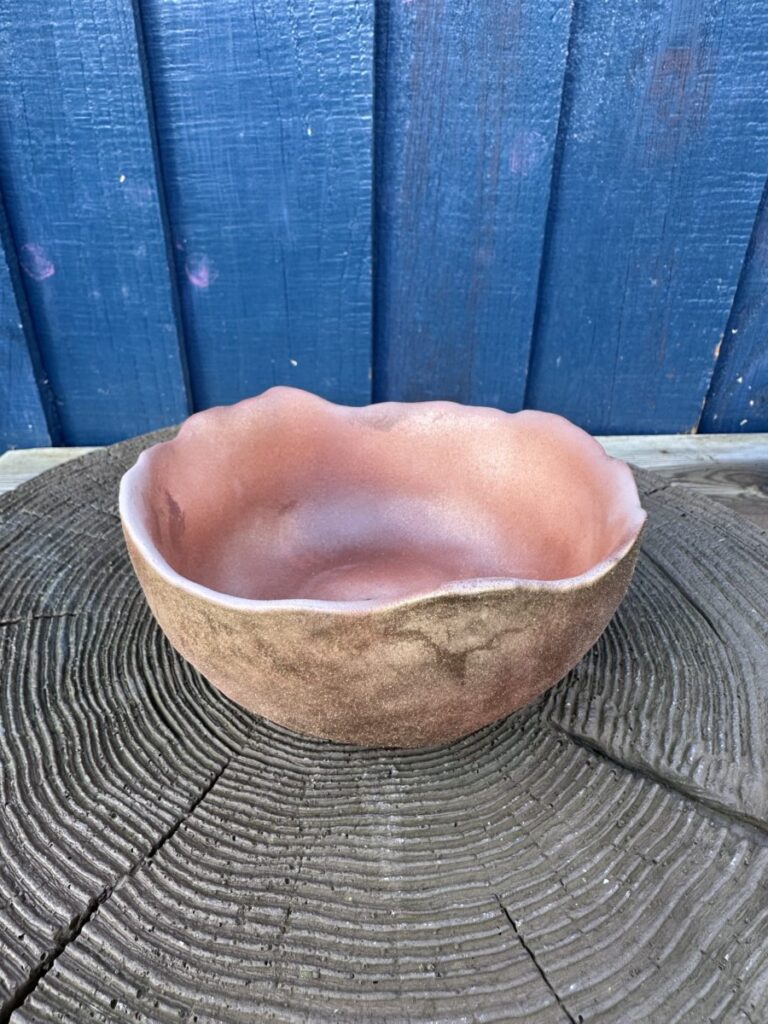 A wide, shallow hand-cast planter bowl with a rustic terracotta color and an irregular, wavy rim, displayed on a textured dark wood surface against a blue wooden backdrop.