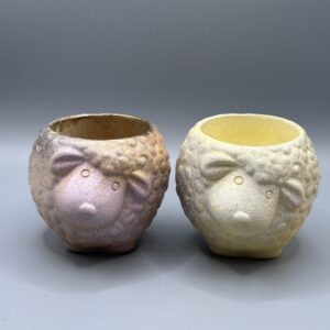 Two small, round sheep-shaped planters with a nubby wool texture; one is finished in a metallic pink and gold ombré, the other in a soft matte yellow.