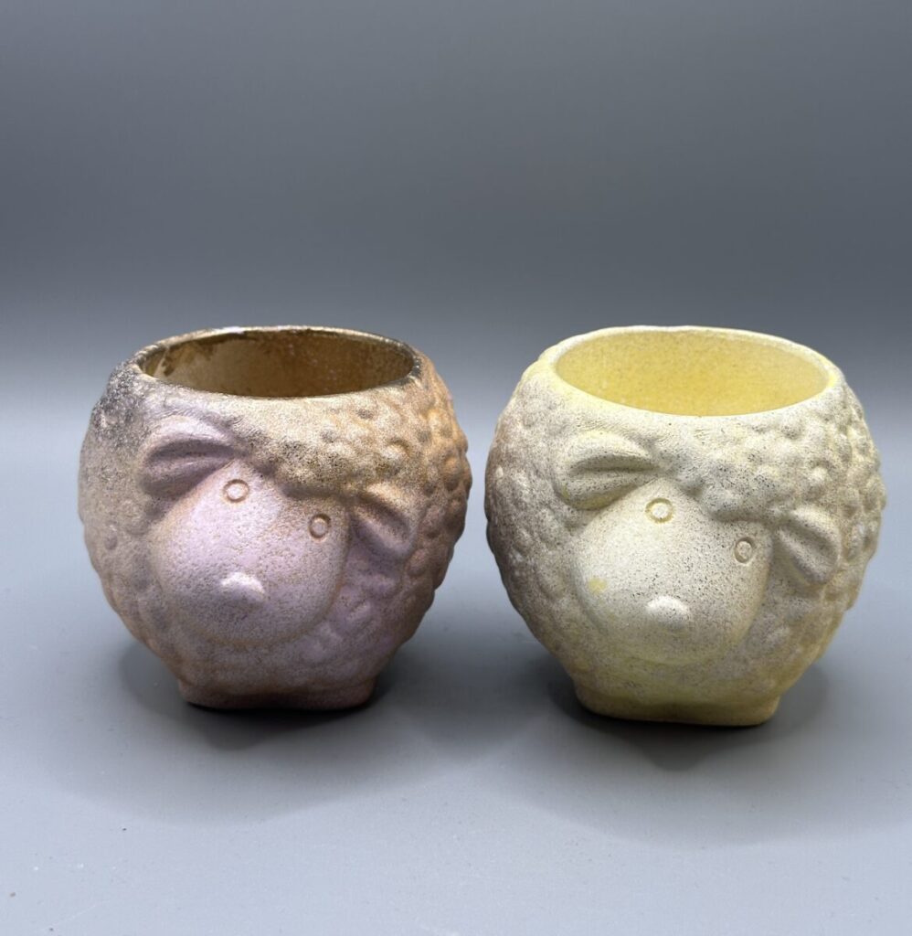 Two small, round sheep-shaped planters with a nubby wool texture; one is finished in a metallic pink and gold ombré, the other in a soft matte yellow.