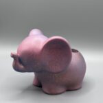 A side view of a small, matte mauve elephant planter with blue and copper speckles, standing against a clean grey background.