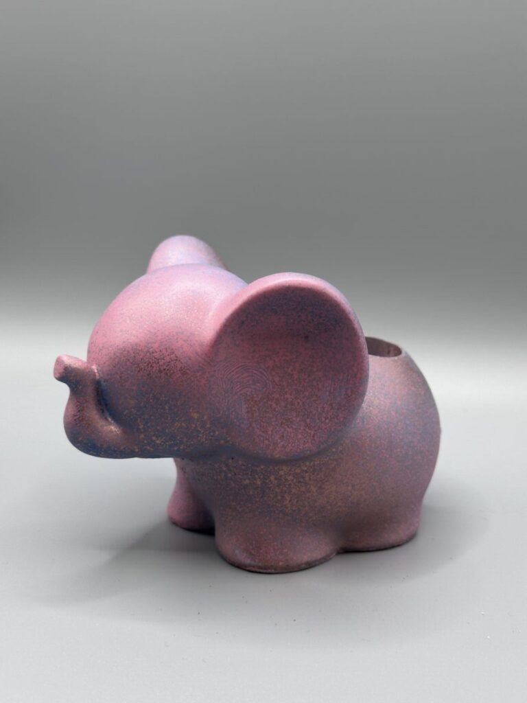 A side view of a small, matte mauve elephant planter with blue and copper speckles, standing against a clean grey background.