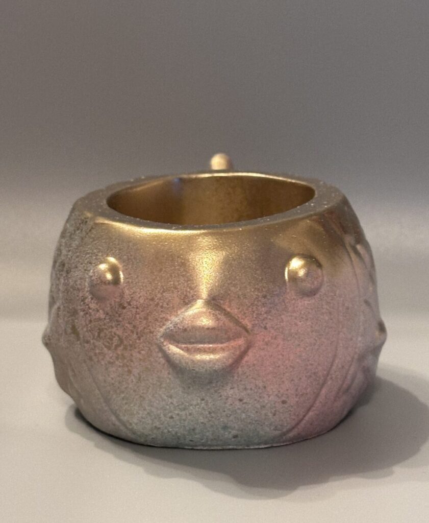 A side profile of a round fish-shaped planter with a metallic gold face transitioning into rose and lilac scales, featuring a sparkling sand-like texture.