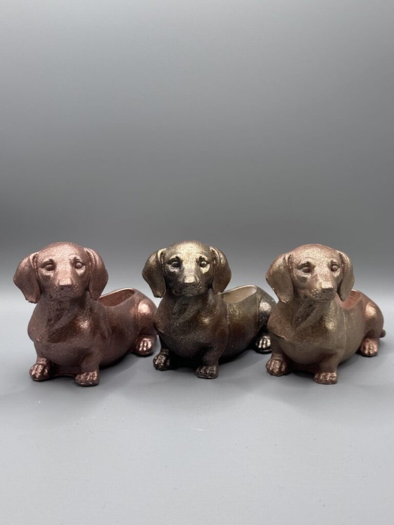 Three small dachshund-shaped planters lined up, featuring textured metallic finishes in rose gold, dark bronze, and warm copper.