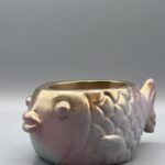 A side profile of a round fish-shaped planter with a metallic gold face transitioning into rose and lilac scales, featuring a sparkling sand-like texture.