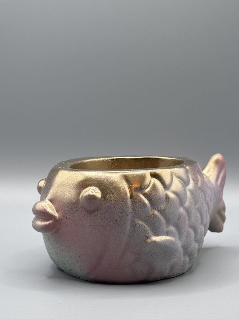 A side profile of a round fish-shaped planter with a metallic gold face transitioning into rose and lilac scales, featuring a sparkling sand-like texture.