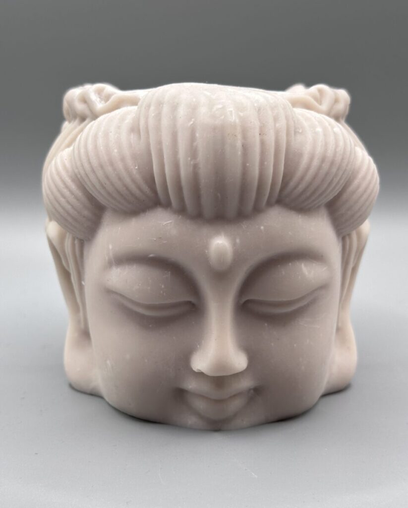 A close-up of a sculptural plant pot shaped like a serene face with closed eyes and detailed swept-back hair, featuring a smooth matte off-white finish against a grey background.