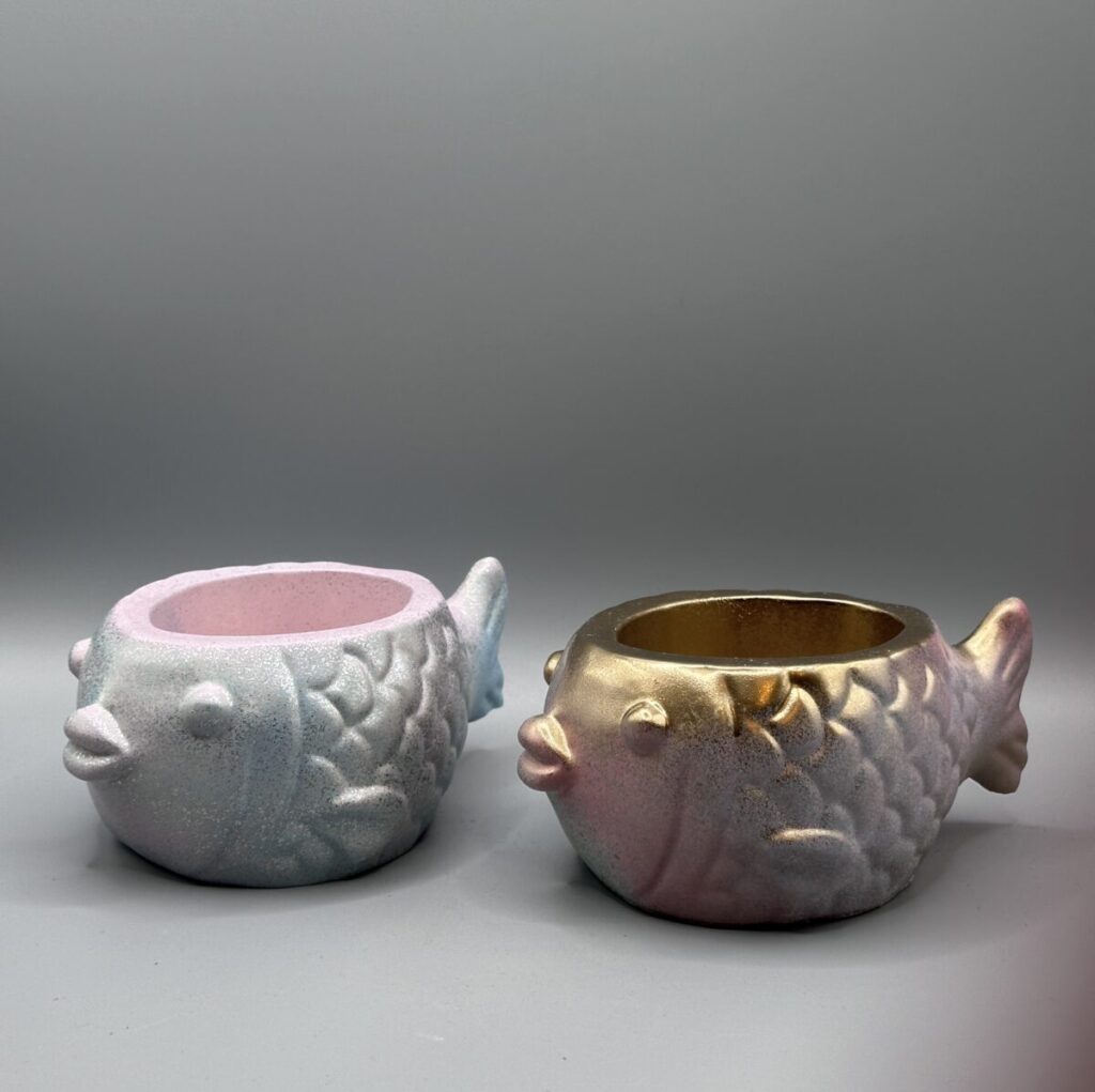 Two small, round fish-shaped planters with embossed scales; one is a shimmering pastel blue and pink, the other is a metallic gold and rose-purple, both set against a clean gray background.