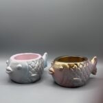 Two small, round fish-shaped planters with embossed scales; one is a shimmering pastel blue and pink, the other is a metallic gold and rose-purple, both set against a clean gray background.