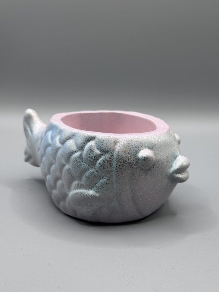 Two small, round fish-shaped planters with embossed scales; one is a shimmering pastel blue and pink, the other is a metallic gold and rose-purple, both set against a clean gray background.