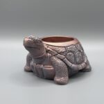 A detailed tortoise-shaped planter with a shimmering metallic bronze and pink finish, featuring a textured shell and friendly expression, set against a grey background.