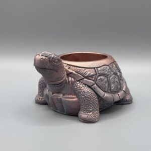 A detailed tortoise-shaped planter with a shimmering metallic bronze and pink finish, featuring a textured shell and friendly expression, set against a grey background.