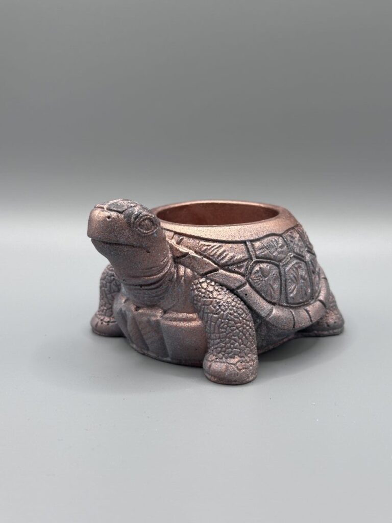 A detailed tortoise-shaped planter with a shimmering metallic bronze and pink finish, featuring a textured shell and friendly expression, set against a grey background.