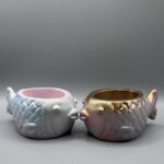 Two small, round fish-shaped planters with embossed scales; one is a shimmering pastel blue and pink, the other is a metallic gold and rose-purple, both set against a clean gray background.