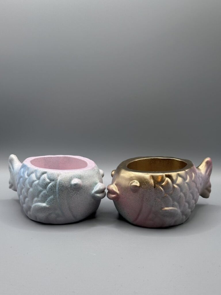 Two small, round fish-shaped planters with embossed scales; one is a shimmering pastel blue and pink, the other is a metallic gold and rose-purple, both set against a clean gray background.