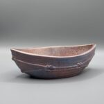 A boat-shaped ceramic vessel with a metallic copper and blue glaze, featuring raised rope textures around the exterior, set against a neutral grey background.