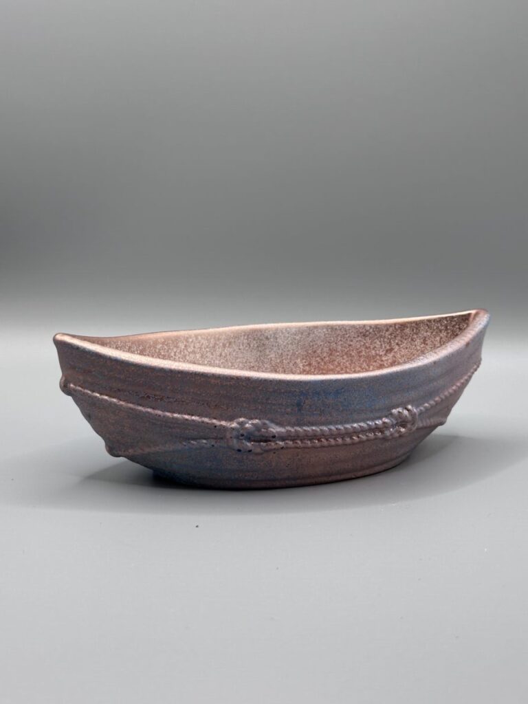 A boat-shaped ceramic vessel with a metallic copper and blue glaze, featuring raised rope textures around the exterior, set against a neutral grey background.