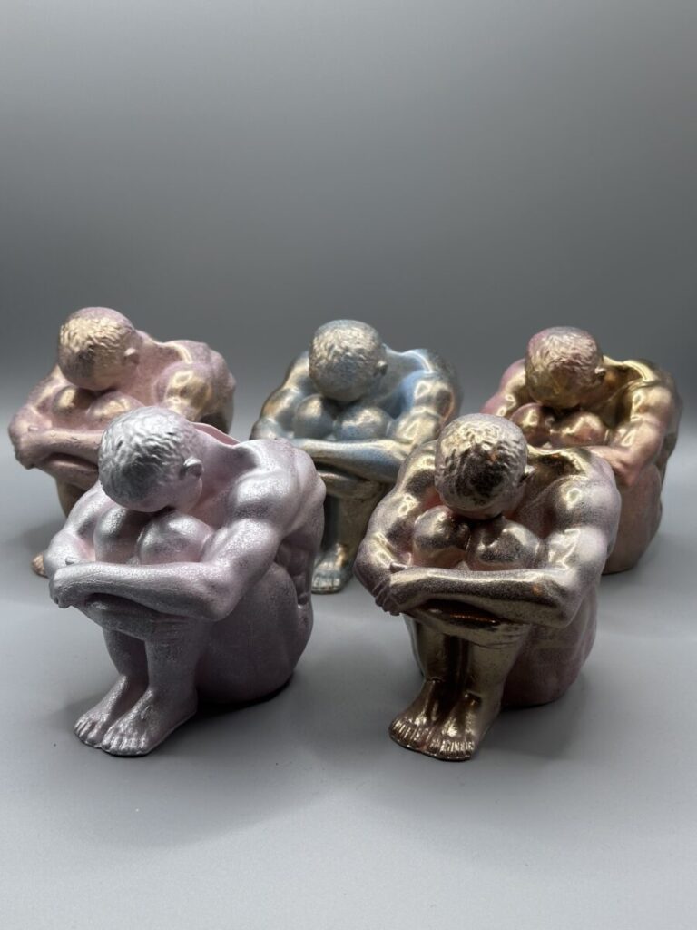 A group of five small sculptural planters shaped like seated, muscular men with bowed heads, featuring various iridescent metallic finishes in silver, gold, pink, and blue.
