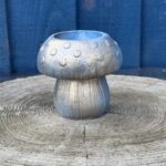 A small hand-cast mushroom-shaped planter with a metallic silver and light blue finish, featuring raised spots on the cap and a ribbed stem, sitting on a light-grey wooden stump.