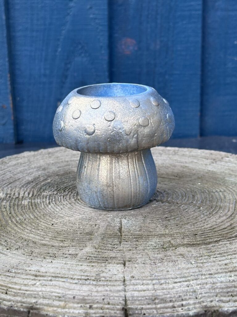 A small hand-cast mushroom-shaped planter with a metallic silver and light blue finish, featuring raised spots on the cap and a ribbed stem, sitting on a light-grey wooden stump.