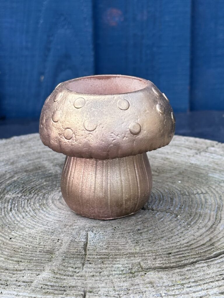 A small, hand-cast planter shaped like a mushroom with a rose gold metallic finish, featuring raised spots on the cap and a ribbed stem, sitting on a light-colored wooden stump.