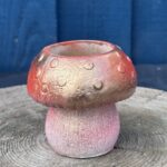 A small hand-cast planter shaped like a mushroom with a gold and red metallic cap and a pink-toned base, sitting on a textured wooden stump against a blue wooden wall.