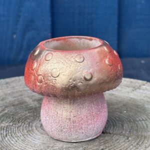 A small hand-cast planter shaped like a mushroom with a gold and red metallic cap and a pink-toned base, sitting on a textured wooden stump against a blue wooden wall.