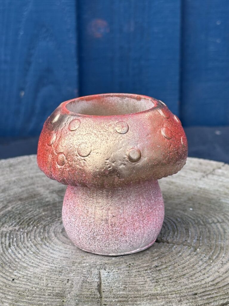 A small hand-cast planter shaped like a mushroom with a gold and red metallic cap and a pink-toned base, sitting on a textured wooden stump against a blue wooden wall.