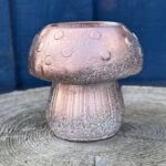 A hand-cast mushroom-shaped planter with a metallic rose gold finish and a white frosted texture at the base, sitting on a weathered wooden surface against a blue wooden background.