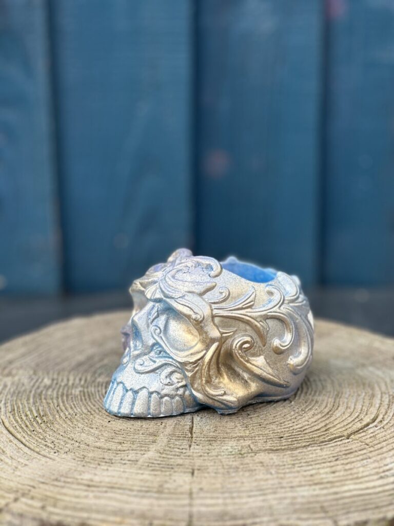 A front-facing view of a highly detailed, ornate sugar skull planter with a shimmering blue and gold iridescent finish, resting on a rustic wood slice against a teal background.