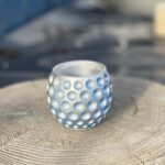 A round, blue dimpled plant pot with silver metallic accents, displayed on a natural wooden tree stump against a blurred blue background.