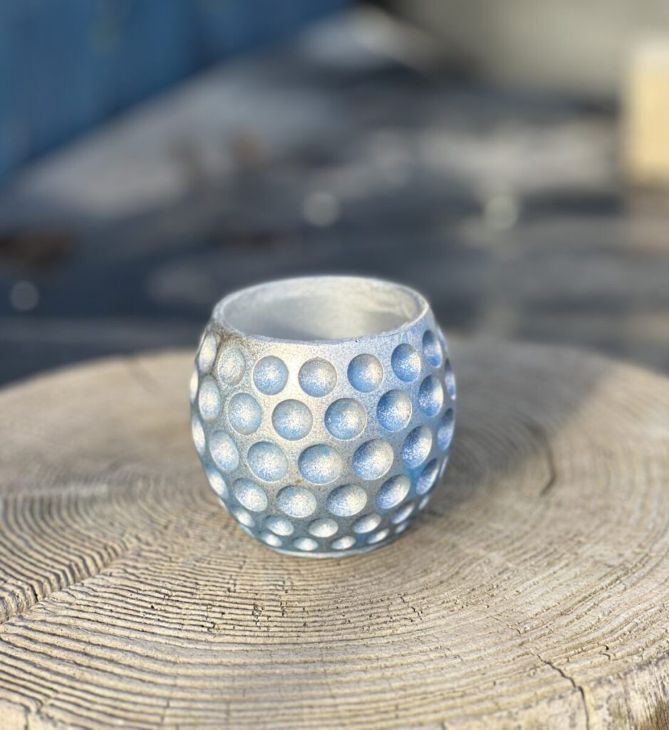 A round, blue dimpled plant pot with silver metallic accents, displayed on a natural wooden tree stump against a blurred blue background.