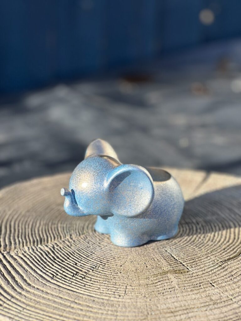 A small, light blue elephant-shaped planter with a shimmering, glittery gold and blue texture sitting on a rustic wooden tree stump.