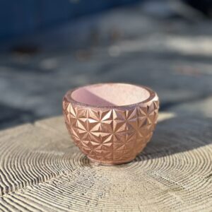 A small, round hand-cast planter with a geometric faceted exterior in a metallic copper color, sitting on a light-colored wooden stump in soft sunlight.