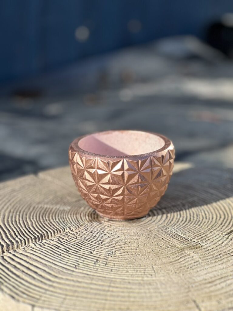 A small, round hand-cast planter with a geometric faceted exterior in a metallic copper color, sitting on a light-colored wooden stump in soft sunlight.
