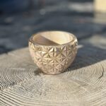 A small, round hand-cast planter with a geometric faceted exterior in a metallic gold color, sitting on a light-colored wooden stump in soft afternoon sunlight.