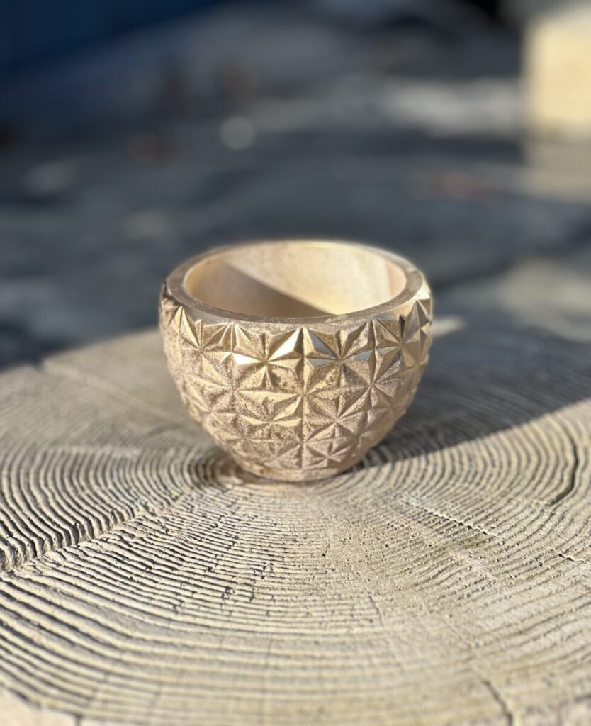 A small, round hand-cast planter with a geometric faceted exterior in a metallic gold color, sitting on a light-colored wooden stump in soft afternoon sunlight.