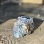 A front-facing view of a highly detailed, ornate sugar skull planter with a shimmering blue and gold iridescent finish, resting on a rustic wood slice against a teal background.