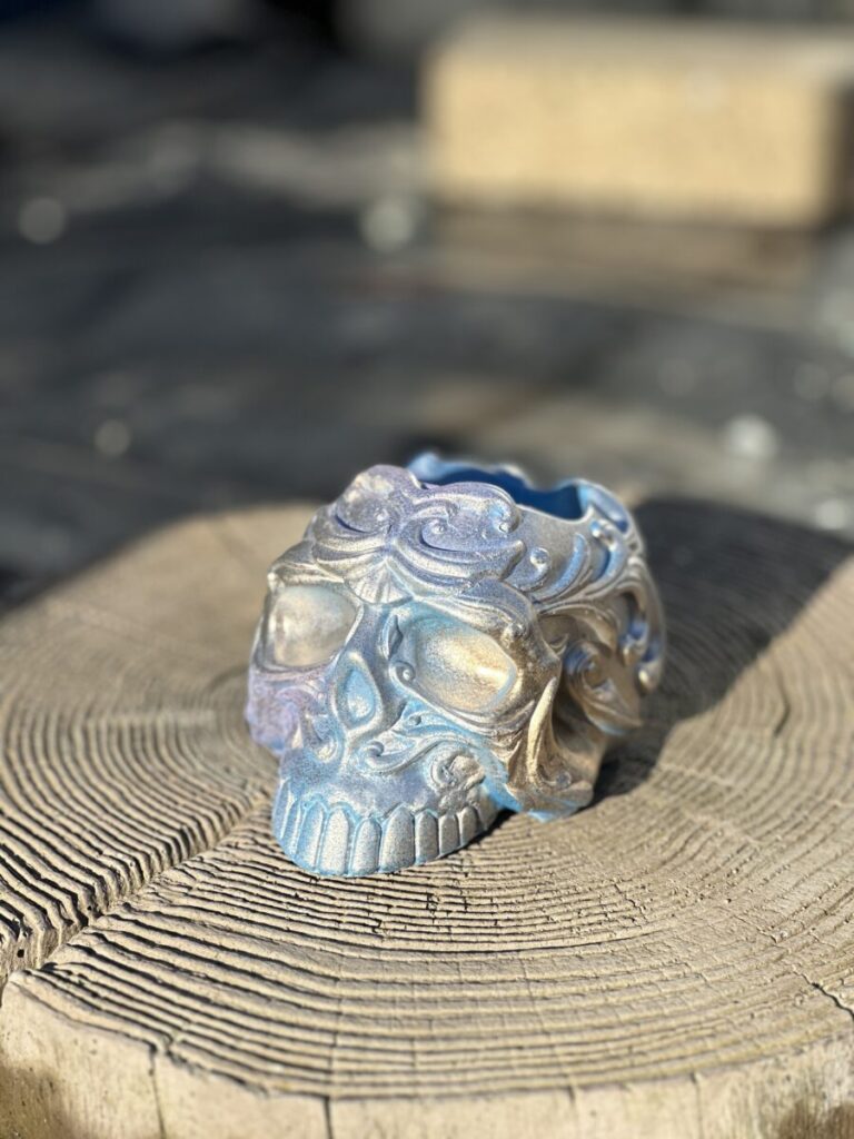 A front-facing view of a highly detailed, ornate sugar skull planter with a shimmering blue and gold iridescent finish, resting on a rustic wood slice against a teal background.