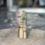 A sculptural face planter with an iridescent purple and sage green color-shift finish, showing a hand holding a metallic rose over one eye, sitting on a natural wood stump.