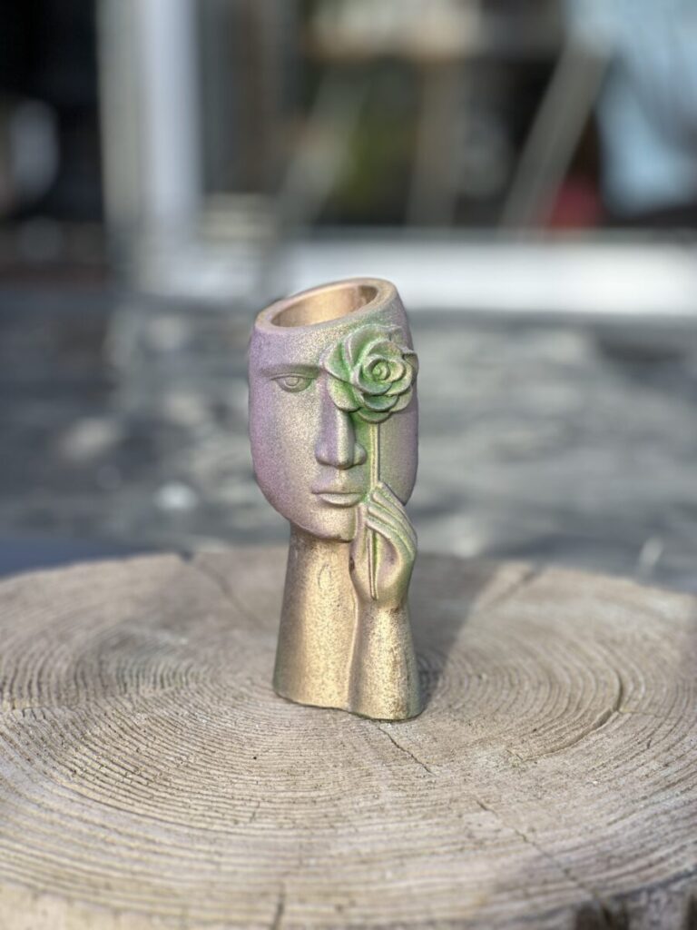 A sculptural face planter with an iridescent purple and sage green color-shift finish, showing a hand holding a metallic rose over one eye, sitting on a natural wood stump.