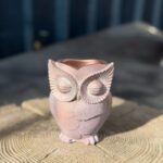 A small, hand-finished owl-shaped planter with a shimmering rose gold stone texture, resting on a rustic wood tree stump.