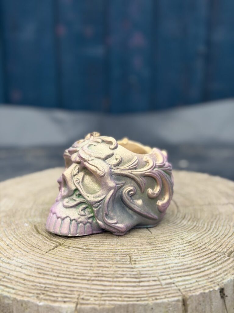 Ornate baroque-style resin skull planter with intricate scrollwork, finished in a chameleon colour-shifting shimmer of dusky pink, olive gold, emerald green, and dark gunmetal, displayed on a natural wood slice in warm sunlight.