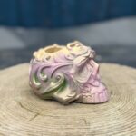 Ornate baroque-style resin skull planter with intricate scrollwork, finished in a chameleon colour-shifting shimmer of dusky pink, olive gold, emerald green, and dark gunmetal, displayed on a natural wood slice in warm sunlight.