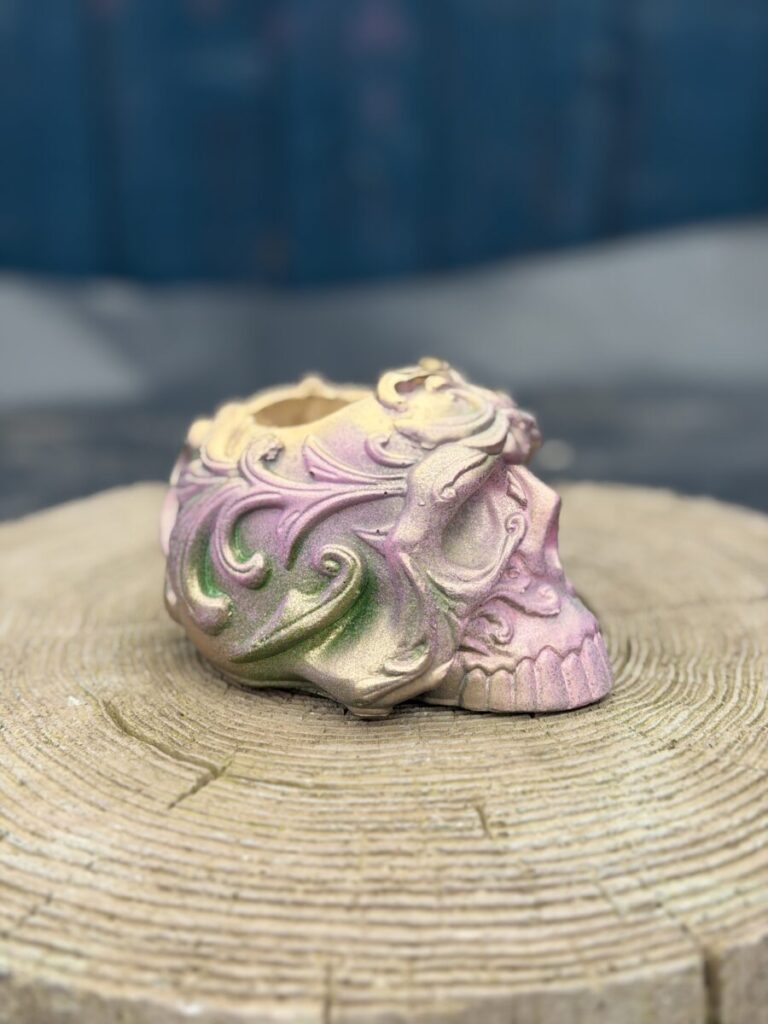 Ornate baroque-style resin skull planter with intricate scrollwork, finished in a chameleon colour-shifting shimmer of dusky pink, olive gold, emerald green, and dark gunmetal, displayed on a natural wood slice in warm sunlight.