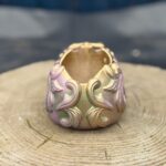 Ornate baroque-style resin skull planter with intricate scrollwork, finished in a chameleon colour-shifting shimmer of dusky pink, olive gold, emerald green, and dark gunmetal, displayed on a natural wood slice in warm sunlight.