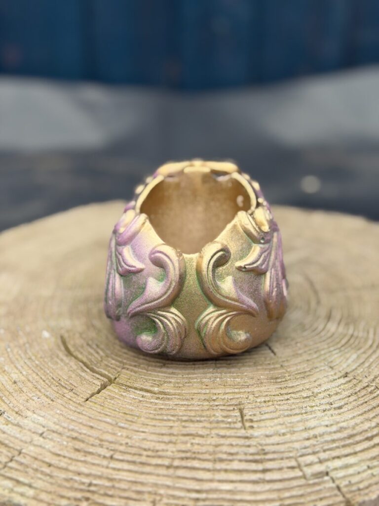 Ornate baroque-style resin skull planter with intricate scrollwork, finished in a chameleon colour-shifting shimmer of dusky pink, olive gold, emerald green, and dark gunmetal, displayed on a natural wood slice in warm sunlight.